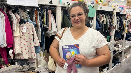 Talea Koch steht lächelnd vor einem Klamottenregal und hält eine Puppe und ein Kinderbuch in der Hand.