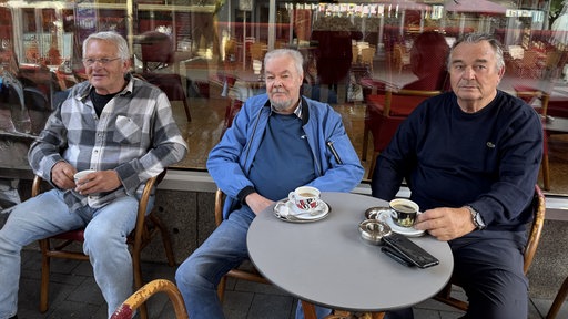 Die drei Männer Manfred, Helmut und Heinz sitzen draußen vor einem Café, vor ihnen stehen Kaffeetassen