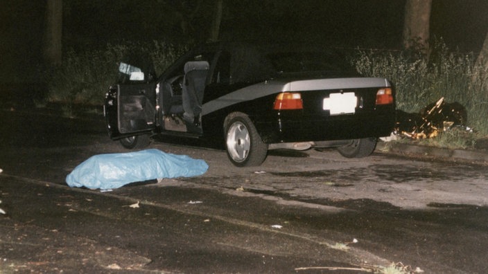 Tatortfoto vom 7. Juni 1999: Ein schwarzes Auto steht mit offener Tür auf der Straße, daneben liegen zwei Leichen, die mit einer blauen und einer goldenen Plane abgedeckt sind
