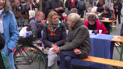 Auf dem Foto sind Menschen, die an einem hölzernen Klapptisch sitzen. Manche von ihnen essen Suppe mit Plastikgabeln.