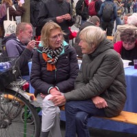 Auf dem Foto sind Menschen, die an einem hölzernen Klapptisch sitzen. Manche von ihnen essen Suppe mit Plastikgabeln.