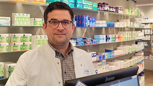 Auf dem Foto ist Felix Tenbieg im weißen Apothekerkittel. Vor ihm ein PC. Hinter ihm Medikamente.