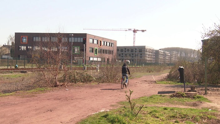 Die Aussicht vom ehemaligen Standort, man sieht Wohnhäuser sowie ein Schulungsstandort, ein Fahrradfahrer fährt auf einem Weg neben den Bahngleisen.