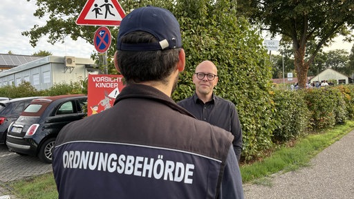Zwei Männer stehen sich gegenüber, der eine trägt Glatze und eine Brille, der andere eine Mütze und eine Weste mit der Aufschrift "Ordnungsbehörde"