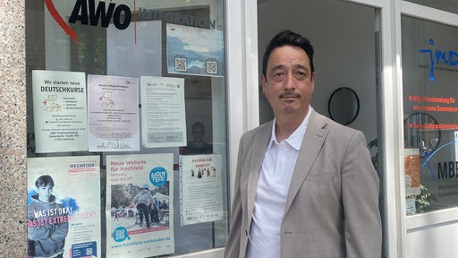 Mike Kim ist Prokurist der AWO-Integration. Er trägt ein hellbraunes Sakko und steht neben einer Fensterscheibe, an der verschiedene Ausdrucke hängen.