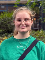 Eine junge Frau mit zusammengebunden blonden Haaren, Brille und einem grünen Pullover lächelt frontal in die Kamera
