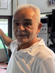 Ein Mann mit kurzen Haaren und weißem Oberteil steht neben einem Dönerspieß und schaut in die Kamera.