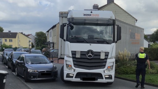 Ein LKW steht in einer engen Wohngebietsstraße quer, daneben steht ein Polizist.