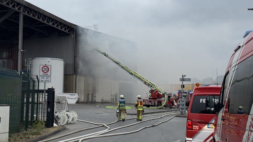 Feuerwehrkräfte und eine ausgefahrene Drehleiter sind vor einem Gebäude zu sehen, dass brannte. Daher sieht man im Hintergrund auch eine Rauchwolke.
