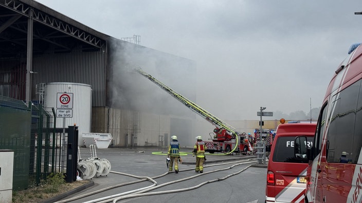 Feuerwehrkräfte und eine ausgefahrene Drehleiter sind vor einem Gebäude zu sehen, dass brannte. Daher sieht man im Hintergrund auch eine Rauchwolke.