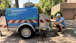Ein Trinkwasserwagen. Davor sitzt ein Mann im blauen Oberteil und blauer Kappe und zapft sich Wasser ab.