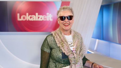 Eine Frau mit kurzen Haaren und Sonnenbrille lächelt in die Kamera. lm Hintergrund steht 'Lokalzeit' auf einem großen Monitor.