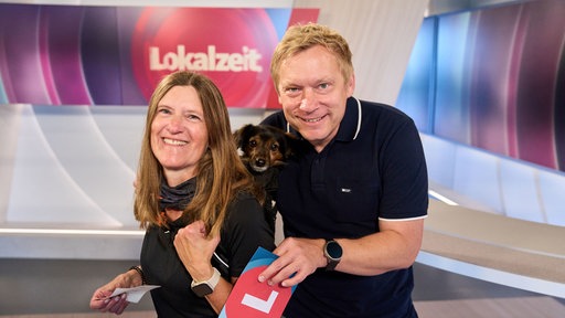 Links eine Frau mit braunen Haaren und einem schwarzen Oberteil. Auf ihrem Rücken trägt sie einen kleinen Hund in einem Rucksack. Rechts daneben ein blonder Mann mit einem dunklen Oberteil. Im Hintergrund ein Monitor, auf dem 'Lokalzeit' steht