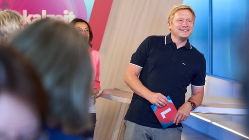 Rechts ein Mann mit hellblonden Haaren und dunklem Oberteil. Er lehnt an einem Tisch und lacht. Links sieht man verschwommen Köpfe von hinten
