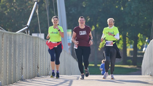 Drei Frauen laufen auf einer Brücke und lächeln in die Kamera. Die linke und die rechte tragen ein neongelbes Oberteil, die Frau in der Mitte ein dunkelrotes.