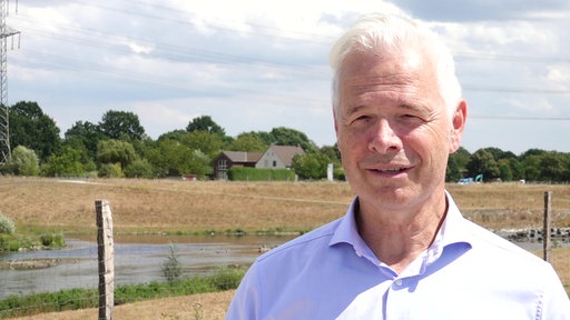 Ein Mann mit weißem Haar und fliederfarbenem Hemd schaut in die Kamera. Im Hintergrund Landschaft und Häuser.