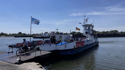 Eine Fähre steht im Wasser an einem Anleger. Zwei Personen verlassen die Fähre auf Fahrrädern