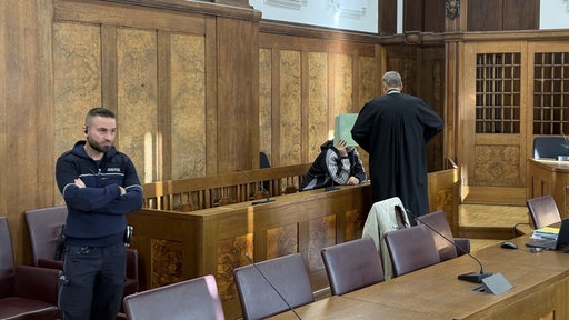 In einem Gerichtssaal sitzt ein Mann, der sich hinter einer grünen Mappe verscteckt. Vor ihm steht eine Person in einer schwarzen Robe. Daneben steht ein dunkelhaariger Mann von der Justiz