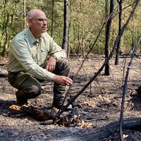 Ein Mann in Försterkleidung hockt an einer verbrannten Waldstelle