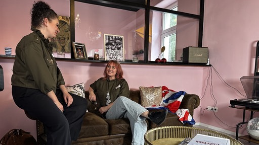 In der Mitte des Bildes sitzt eine rothaarige Frau auf einer Couch, links von ihr sitzt eine schwarzhaarige Frau auf der Armlehne. Die beiden schauen sich an.