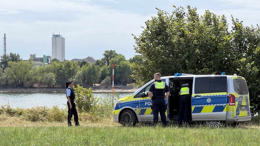 Eine Wiese. Links steht eine Polizistin, rechts ein Polizist vor einem Polizeiauto. Im Hintergrund der Rhein. 