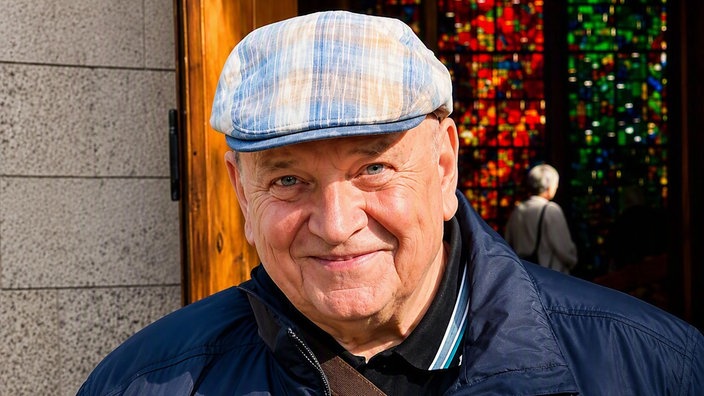 Ein älterer Mann mit Mütze und blauer Jacke lächelt freundlich in die Kamera