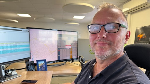Ein Mann mit leichtem grauen Bart und Brille sitzt an einem Schreibtisch vor einem Computer und lächelt in die Kamera