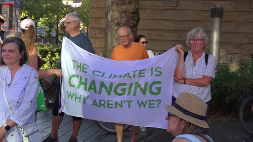 Drei Menschen, zwei Männer und eine Frau, halten eine weiß-grüne Fahne mit der Aufschrift: "The climate is changing why aren't we?"