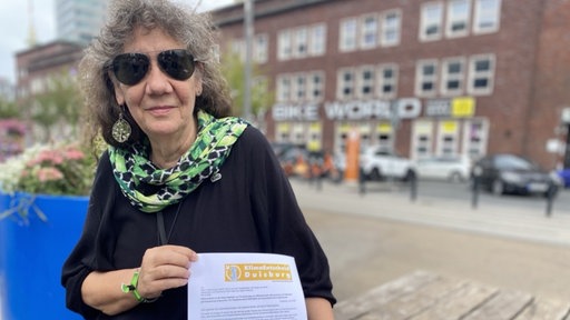 Eine Frau mit grauen Haaren, einem dunklen Oberteil und einem grünen Halstuch hält einen Zettel in beiden Händen