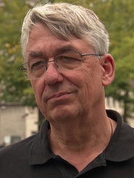 Ein Mann mit hellen Haaren und Brille schaut in die Kamera. Er trägt ein schwarzes Shirt. Im Hintergrund die Duisburger Innenstadt