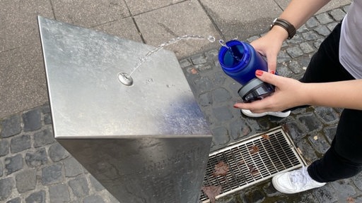 Ein Trinkwasserbrunnen von oben fotografiert. Links daneben hält jemand eine blaue Trinkwasserflasche und das Wasser vom Brunnen fließt dort hinein.