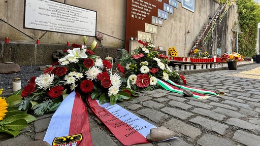 Blumenkränze und Grabkerzen stehen vor einer Wand, im Hintergrund sind an der Wand außerdem Fotos und Texte sowie eine Steintreppe zu sehen
