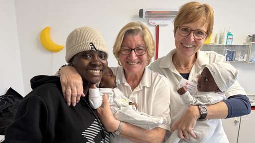 Dr. Delia Streuter und Hebamme Doris Kammann mit ihrer früheren Patientin Mariama. Die drei Frauen lächeln in die Kamera und halten zwei Babys.