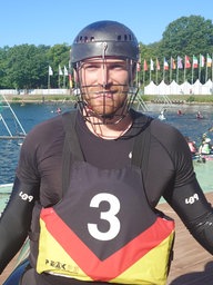 Ein Mann mit einem schwarzen Neoprenanzug mit der Nummer drei und einem Helm mit Schutz vor dem Gesicht guckt in die Kamera