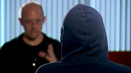 Ein Insider berichtet von den Vorwürfen, ist nur von hinten zu sehen, trägt zur Anonymisierung einen Kaputzenpullover, im Hintergrund ist der Reporter unscharf zu sehen.