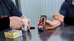 Zwei unkenntlich gemachte Personen sitzen an einem Tisch und zeigen eine Bild auf einem Handy