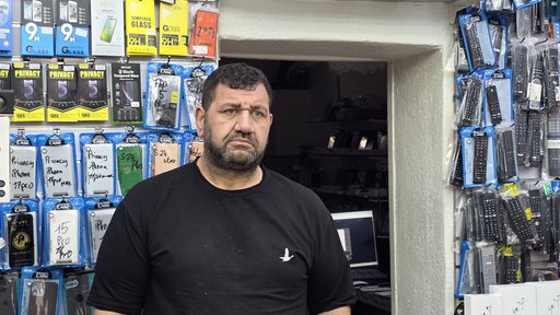 Ladenbesitzer Mehmet Sönmez trägt ein schwarzes Shirt und hinter ihm hängt buntes Handyzubehör an der Wand.