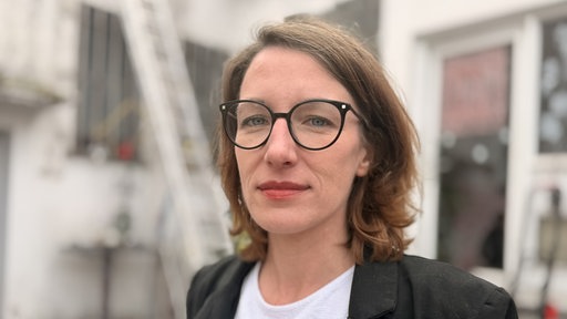 Lena Wiese (Verein für die solidarische Gesellschaft der Vielen e.V.) trägt eine schwarze Brille und ein schwarz-weißes Outfit.