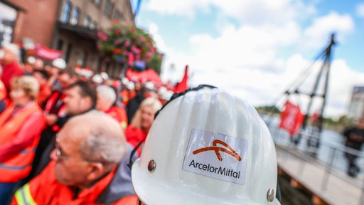 Ein Helm mit der aufschrift "ArcelorMittal" ist im Vordergrund und im Hintergrund mehrere menschen mit Warnwesten.