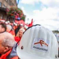 Ein Helm mit der Aufschrift "ArcelorMittal" ist im Vordergrund und im Hintergrund mehrere Menschen in Warnwesten.