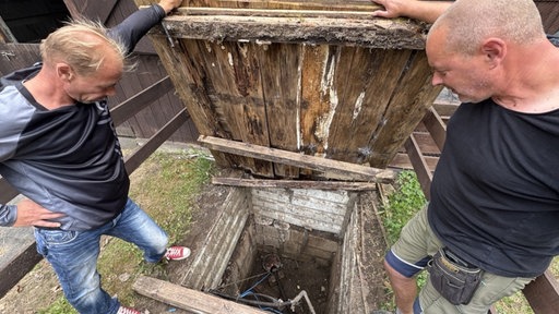 Zwei Männer in dunklen Oberteilen und Jeans stehen über einen leeren Brunnen