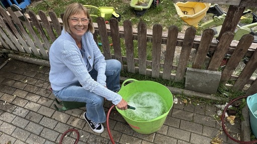 Eine Frau mit einer blauen Strickjacke sitzt auf einem Hocker und füllt mit einem Gartenschlauch Wasser in einen Eimer