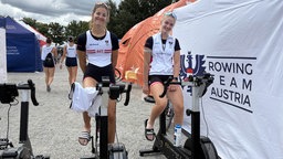 Zwei Frauen in Sportkleidung sitzen auf Trainingsfahrrädern und lächeln in die Kamera.