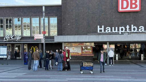 Eine Gruppe Menschen steht für einem Großen Gebäude. Neben ihnen steht ein kleiner blauer Wagen mit einem Poster auf dem steht "Die Reichen wollen Krieg".