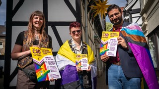 Drei junge Menschen gucken in die Kamera und halten demonstrativ Plakate, die auf den ersten CSD in Hattingen hinweisen. Zwei Frauen und ein Mann. Zwei von ihnen tragen Regenbogenfahnen als Umhang.