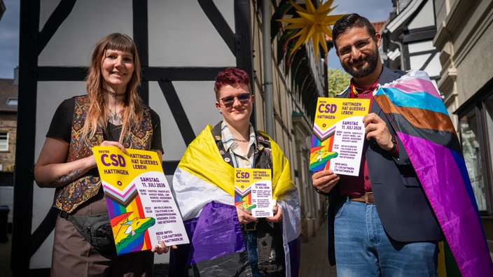 Drei junge Menschen gucken in die Kamera und halten demonstrativ Plakate, die auf den ersten CSD in Hattingen hinweisen. Zwei Frauen und ein Mann. Zwei von ihnen tragen Regenbogenfahnen als Umhang.