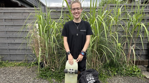 Mia Djuric hat ihr Skateboard und einen Rucksack dabei und lächelt in die Kamera.