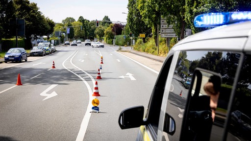 Ein Polizeiauto fährt eine Straße herunter. Auf der Straße stehen mehrere rot-weiß-gestreifte Pylonen. An der Seite parken Autos. Die Scheibe an der Seite des Fahrers bei dem Polizeiauto ist geöffnet. Innen sieht man einen Polizisten sitzen. 