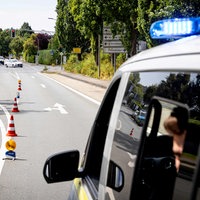 Ein Polizeiauto fährt eine Straße herunter. Auf der Straße stehen mehrere rot-weiß-gestreifte Pylonen. An der Seite parken Autos. Die Scheibe an der Seite des Fahrers bei dem Polizeiauto ist geöffnet. Innen sieht man einen Polizisten sitzen. 