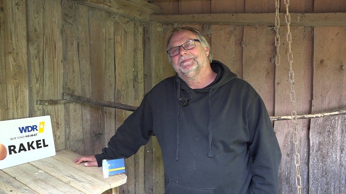 Ein Mann mit Brille und schwarzem Pullover steht in einem Hühnerstall.